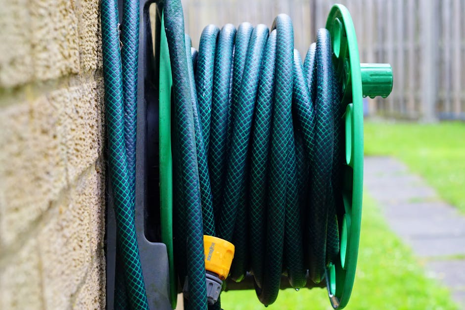 A close-up view of a garden hose reel mounted on an outdoor wall, featuring a black and green plastic frame and a coiled blue garden hose with a diamond pattern. The hose is neatly wound around the re