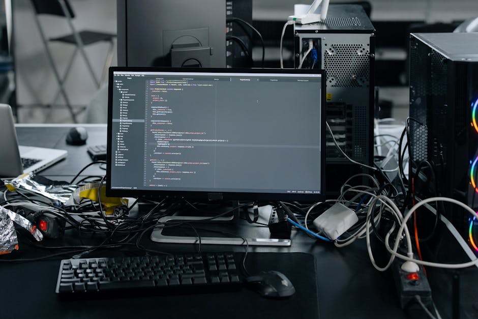 A silver laptop computer with a black keyboard is open on a wooden desk in a well-lit room. The screen displays lines of code in a text editor, with visible syntax highlighting and a sidebar showing p